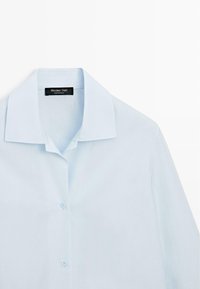 Lichtblauw overhemd met kraag en knopen, met een zwart "Massimo Dutti" label genaaid aan de binnenkant van de kraag, plat neergelegd op een witte achtergrond.