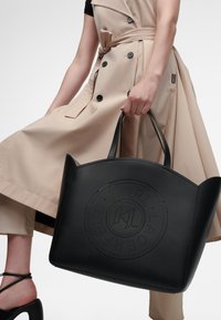 Sac fourre-tout noir en cuir, avec un design de logo perforé, une forme structurée et des doubles poignées. Le modèle porte un trench-coat beige.
