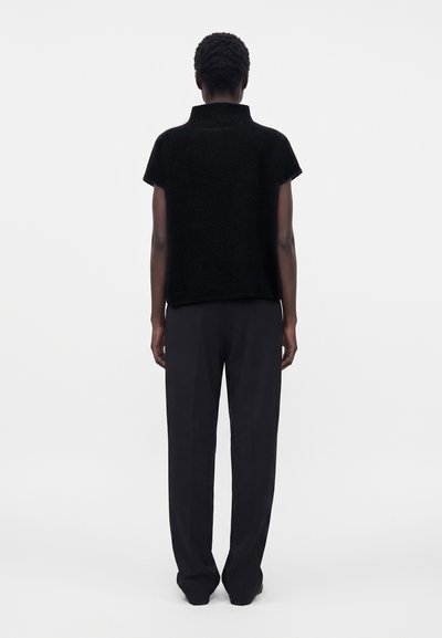Personne debout, de dos, portant un haut noir à manches courtes avec un col montant et un pantalon noir ample sur un fond uni.