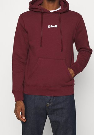 Maroonfarbener Hoodie mit Fronttasche und weißem "Schott"-Logo mittig auf der Brust, getragen von einer Person mit einer Hand in der Tasche und dunkelblauen Jeans.