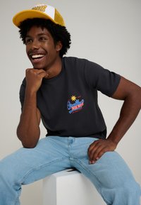 Black T-shirt with colorful "Bradley's Big Buy" logo, light blue jeans, and a yellow mesh cap with white front panel. Smooth fabric, relaxed fit.