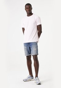 T-shirt en coton rose clair, shorts en jean avec bords effilochés et manches retroussées, baskets grises avec accents bleus. Design simple et décontracté.
