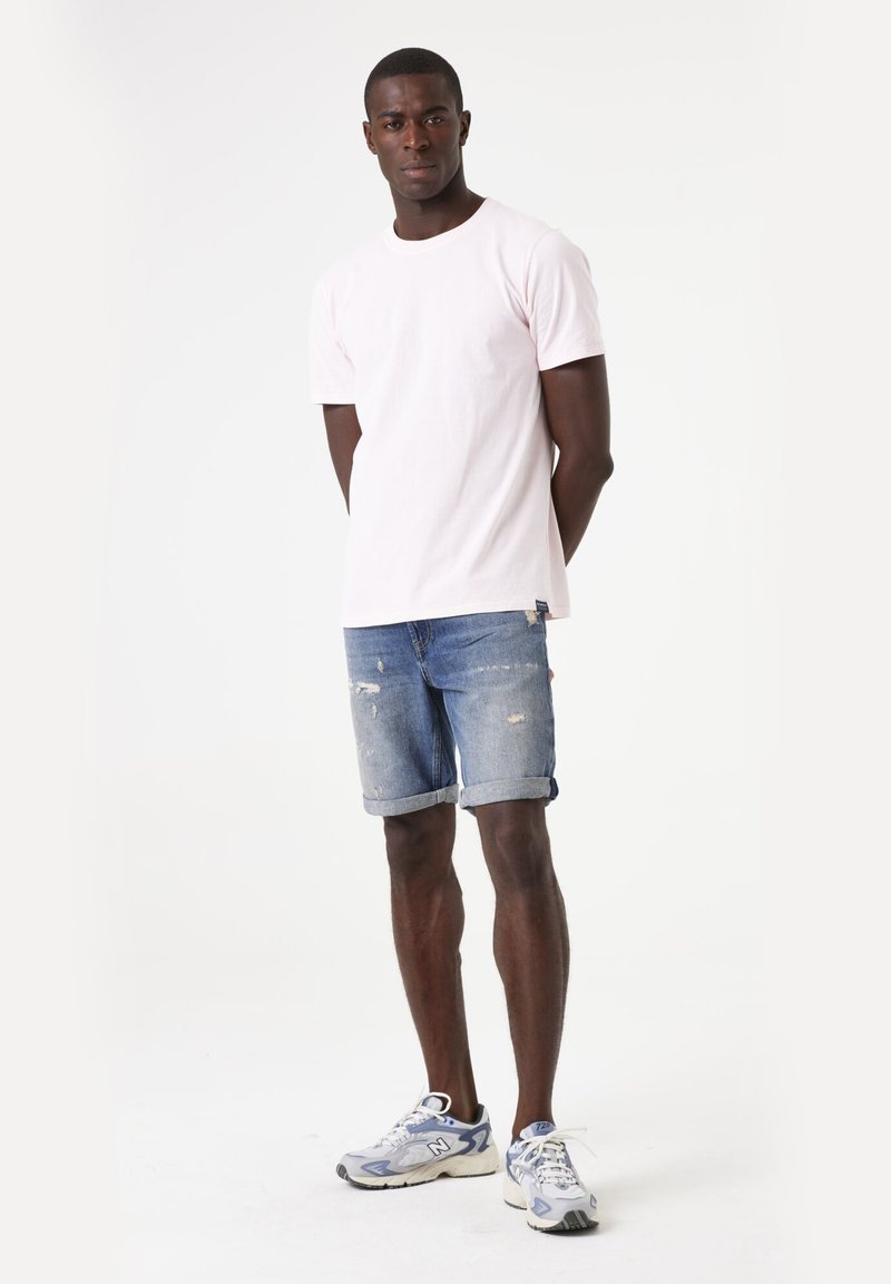 T-shirt en coton rose clair, shorts en jean avec bords effilochés et manches retroussées, baskets grises avec accents bleus. Design simple et décontracté.