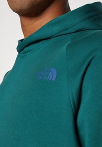 Felpa con cappuccio in misto cotone teal, caratterizzata da una finitura texture e un logo ricamato blu sul lato sinistro del petto.