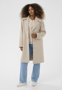 Cappotto in misto lana beige con colletto a rever, bottoni frontali e tasche laterali, indossato sopra un top beige chiaro e jeans a gamba larga blu.
