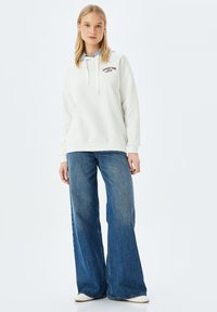 Sudadera blanca con "SUPPORT YOUR" bordado en negro, combinada con jeans de mezclilla azul de pierna ancha. El conjunto presenta un diseño casual y un ajuste relajado.