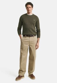 Maglione verde a treccia, pantaloni beige e scarpe marroni con suola spessa. L'outfit presenta un design classico con una vestibilità comoda.