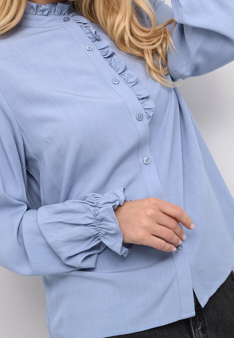 Blouse bleu clair avec col et poignets à volants, boutonnée devant, portée par une personne aux cheveux blonds et au teint clair.