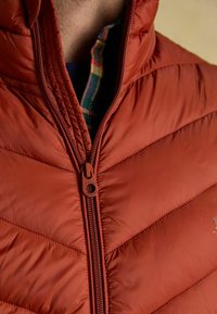 Červená prešívaná pufrová vesta s vysokým golierom, predným zipsom a so značkovým ťahadlom na zips. Pod ňou je vidieť károvanú košeľu.