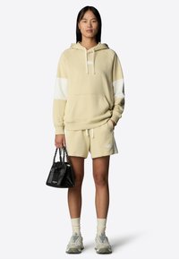 Felpa beige con accenti bianchi, pantaloni corti coordinati e una piccola borsa nera. Tessuto texturizzato, cappuccio con coulisse e dettaglio del logo.
