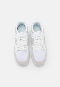 Pár bílých atletických tenisek s béžovými semišovými detaily, bílými tkaničkami a prodyšnou síťovinou, viděný zhora na jednobarevném pozadí.