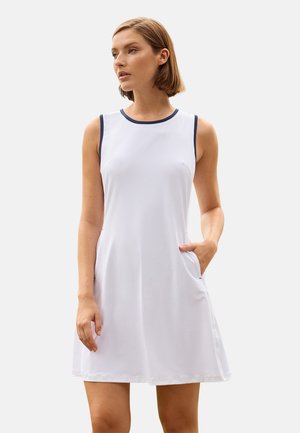 Mujer con vestido blanco sin mangas con ribete azul marino, de pie con una mano en el bolsillo, mirando ligeramente hacia un lado sobre un fondo liso.
