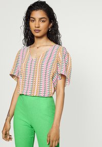 Top multicolor com decote em V, mangas curtas e padrões geométricos em rosa, amarelo e azul. Combinado com calças de cintura alta verdes com nervuras.