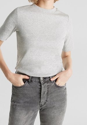 Person trägt ein hellgraues, kurzärmeliges Hemd, das in eine hoch taillierte, ausgeblichene schwarze Jeans gesteckt ist, mit den Händen in den Vorderhosentaschen.