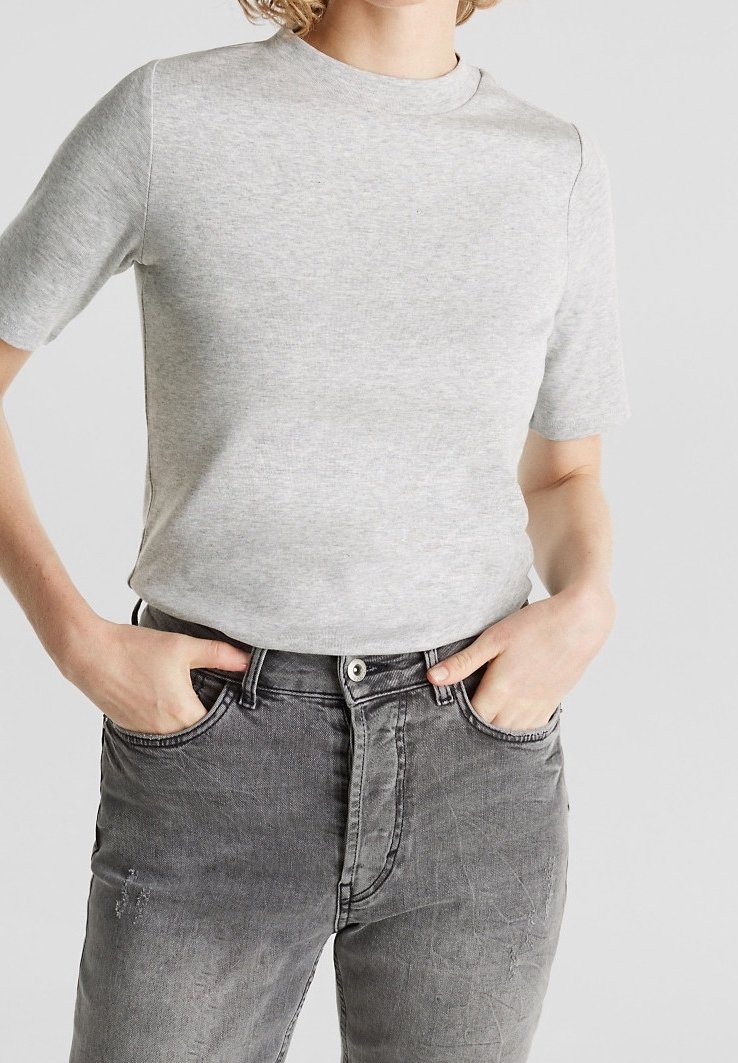 Personne portant une chemise gris clair à manches courtes rentrée dans un jean noir délavé taille haute, les mains dans les poches avant.