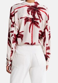 Camicia bianca a maniche lunghe con stampa di palme rosse. Tessuto strutturato, vestibilità rilassata, polsini con bottoni e dettaglio sulla parte posteriore. Abbinata a pantaloni bianchi.