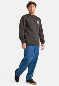 Sudadera gris oscuro con puños y cuello acanalados, con un pequeño parche gráfico. Combinada con pantalones de mezclilla azul sueltos y zapatillas negras.