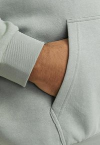 Lys grå sweatshirt med ribbet manchet og sidelomme, med et nærbillede af en hånd, der hviler i lommen mod et struktureret stof.