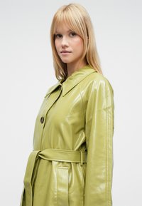 Lysegrønn, blank lær trenchcoat med belte i livet, stor krage og knappestenging. Har sidelommer.