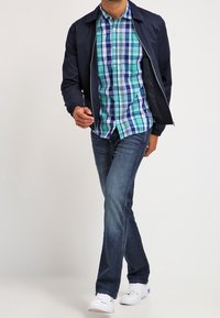 Man som går klädd i en marinblå jacka, blå- och grönrutig skjorta, mörkblå jeans och vita sneakers mot en vit bakgrund.