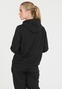 Nainen, jolla on vaalea poninhäntä, pukeutuneena mustaan huppariin ja mustiin housuihin, katsoo vasemmalle yksinkertaista valkoista taustaa vasten.