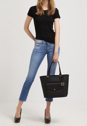 Bolso tote de cuero negro con un bolsillo frontal, herrajes dorados y un charm con el logo. Tiene dos asas y una forma rectangular estructurada.
