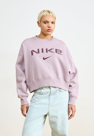 Pessoa com cabelo loiro curto, vestindo um sweatshirt lilás da Nike e umas calças de ganga azul claro, em frente a um fundo branco simples.