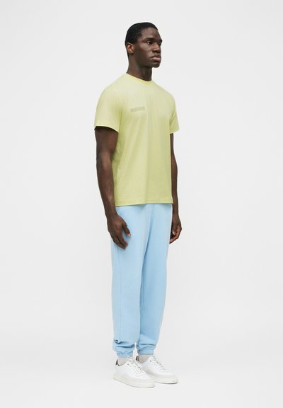 Άνδρας που στέκεται και κοιτά προς τα δεξιά, φοράει ανοιχτόκίτρινη κοντομάνικη μπλούζα, ανοιχτό μπλε παντελόνι τζόγκινγκ και λευκά παπούτσια, σε λευκό φόντο.