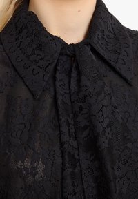 Schwarze Spitzenbluse mit Kragen und Bindedetail, versehen mit floralen Mustern und einer strukturierten Oberfläche. Das durchscheinende Material hebt das filigrane Design hervor.