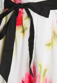 Blommig klänning med vit bas och röda samt gröna mönster, accenterad av ett svart tygbälte som är knutet i en båge vid midjan.