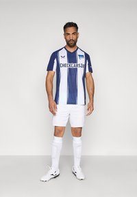 Zila un baltā svītraina futbola krekls ar īsām piedurknēm un V-veida kakla izgriezumu, kombinēts ar baltām šortiem un zeķēm. Satur sponsoru logotipus.