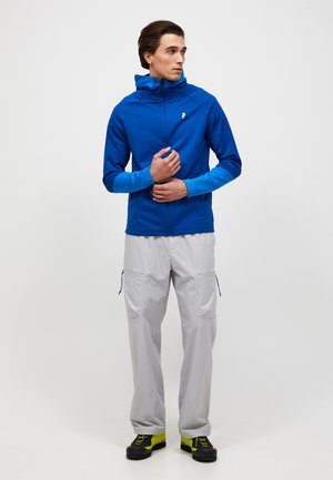 Man draagt een blauw jack met ritssluiting en lichtgrijze cargobroek, staat met samengevouwen handen en kijkt opzij tegen een effen achtergrond.