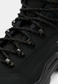 Gros plan d'une chaussure de randonnée noire avec des lacets épais à motifs passés dans des crochets métalliques et une semelle robustes visible sur le côté.
