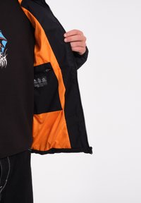 Zwarte jas met oranje voering, met ritssluiting aan de voorkant en een binnenzak. Heeft een gladde textuur en een casual ontwerp met een logo op het shirt dat eronder zichtbaar is.