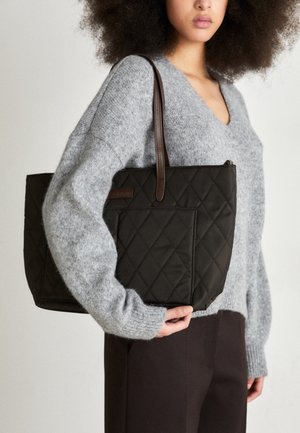 Vrouw met grijze trui en zwarte broek houdt grote zwarte gewatteerde tas met bruine handvatten over de schouder.