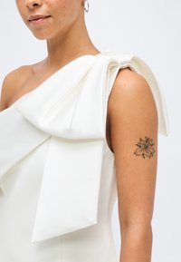 Γυναίκα που φοράει λευκό φόρεμα με έναν ώμο και μεγάλο φιόγκο στον ώμο, με λουλουδένιο τατουάζ στον επάνω βραχίονα.