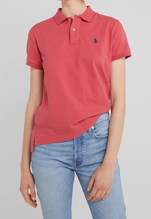 Persona que lleva una camiseta polo roja de manga corta con un pequeño logo azul marino en el pecho y vaqueros azul claro sobre un fondo sencillo.
