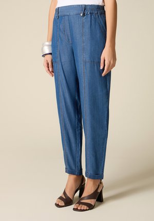 Kobieta nosząca niebieskie dżinsy z wysokim stanem, przednimi kieszeniami, podwiniętymi nogawkami, białą bluzkę, srebrne bransoletki i ciemnobrązowe buty na obcasie z odkrytymi palcami.