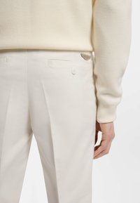 Κοντινό πλάνο ενός ατόμου που φοράει εκρού παντελόνια ραμμένα κατ' ευθείαν, με πίσω τσέπες που κλείνουν με κουμπιά και μικρό λογότυπο της μάρκας πάνω από τη δεξιά τσέπη.