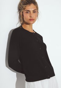 Frau mit blonden Haaren, die eine geknöpfte schwarze Strickjacke und einen weißen Rock trägt, steht mit den Händen hinter dem Rücken gegen eine weiße Wand.