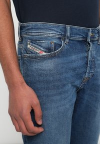 Μπλε τζιν παντελόνι με ευθεία γραμμή, με διακριτικό σχέδιο τσέπης και κόκκινη και λευκή ετικέτα κοντά στην μπροστινή τσέπη. Ορατές ραφές λεπτομερειών.