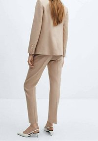 Costume beige sur mesure avec blazer structuré et pantalon slim. Tissu doux, détail à un bouton, associé à des talons blancs à bride arrière.