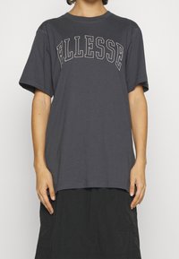 Camiseta gris de manga corta con gran logotipo "ELLESSE" en blanco, cuello redondo, corte relajado y material de algodón suave. Combinada con una falda oscura y fluida.