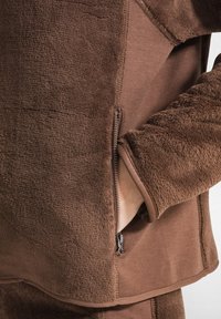 Giacca in pile marrone con una superficie testurizzata, dotata di una tasca laterale con zip e di un rivestimento interno liscio. Le cuciture sono visibili lungo i bordi.