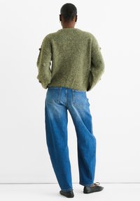Grüner Strickpullover mit strukturierten Ärmeln, kombiniert mit hochgeschnittenen blauen Jeans. Rückansicht, die die lockere Passform und einfache schwarze Schuhe zeigt.