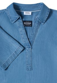 Chemise en denim bleu clair à manches courtes, avec col, ouverture en V, et deux étiquettes visibles portant les mentions "CECIL" et "ocean."