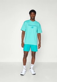 Turkio spalvos medvilniniai marškinėliai su užrašu „Miami Dolphins“ ir logotipu, derinami su atitinkančiais šortais ir baltais sportbačiais su juodais akcentais.