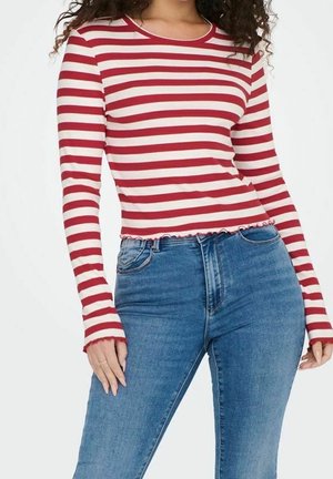 Frau trägt ein rot-weiß gestreiftes Langarmshirt mit gerüschten Kanten und hoch taillierte blaue Jeans, Hände entspannt an den Seiten.