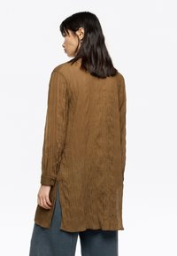 Long, textured brown shirt with vertical pleats, buttoned cuffs, and side slits, designed with a loose fit. Visible seams and soft fabric.