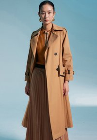 Manteau trench beige avec un col à revers, boutons double boutonnage et poches latérales, superposé sur un haut en maille moutarde et une jupe plissée beige.